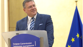 A man in a navy blue suit, light blue shirt, and striped tie stands at a podium labeled "European Commission" in both English and French. He is speaking into a microphone and smiling while looking to the side. A European Union flag is visible to his right against a beige background.